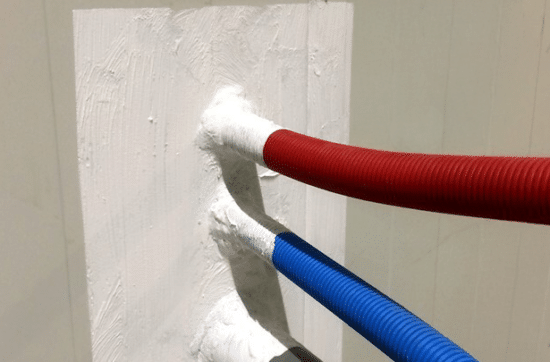 Rode en blauwe ge&iuml;soleerde leidingen die door een brandveilige coating op een muur komen, illustrerend voor brandpreventiemethoden van Grazy.