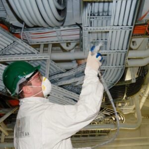 Werknemer in beschermende kleding spuit brandwerende coating op leidingen en kabels in een commerci&euml;le omgeving, benadrukt de toepassing van brandveiligheid in gebouwen.