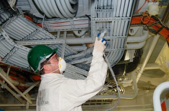 Man in protective gear applying fire-resistant coating to overhead cables in a construction setting, emphasizing fire safety measures.